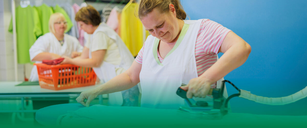 Frau bügelt Wäsche, während zwei weitere Mitarbeiterinnen im Hintergrund Kleidung sortieren